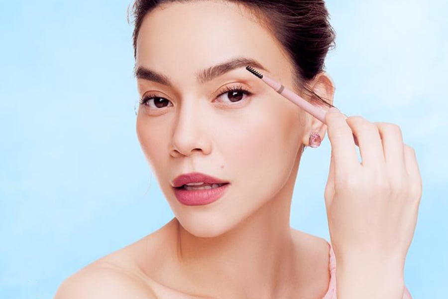 Người phụ nữ trang điểm lông mày bằng cọ, nền trời xanh nhạt phía sau.