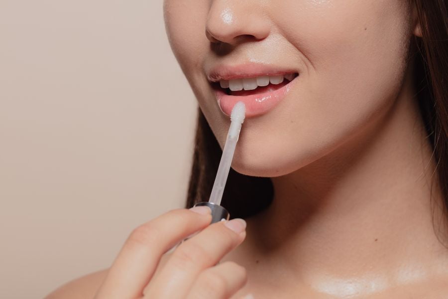 Người cầm cọ thoa son bóng lên môi, cận cảnh nửa khuôn mặt với làn da mịn màng.