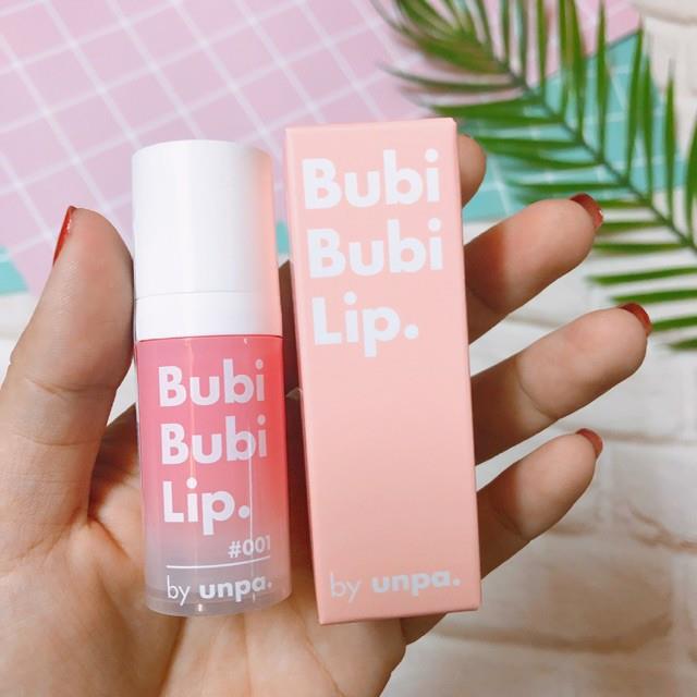 cach-su-dung-bubi-bubi-lip-1 Người cầm sản phẩm dưỡng môi Bubi Bubi Lip màu hồng, nền có lá cây và bàn cờ màu sắc.