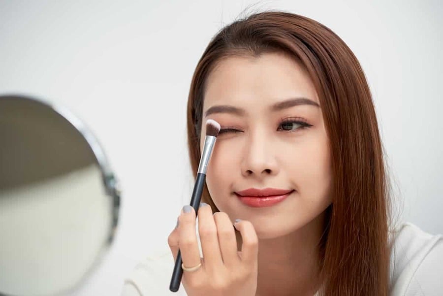 Người phụ nữ cầm cọ trang điểm, đứng trước gương, đang thoa phấn mắt lên mí mắt.