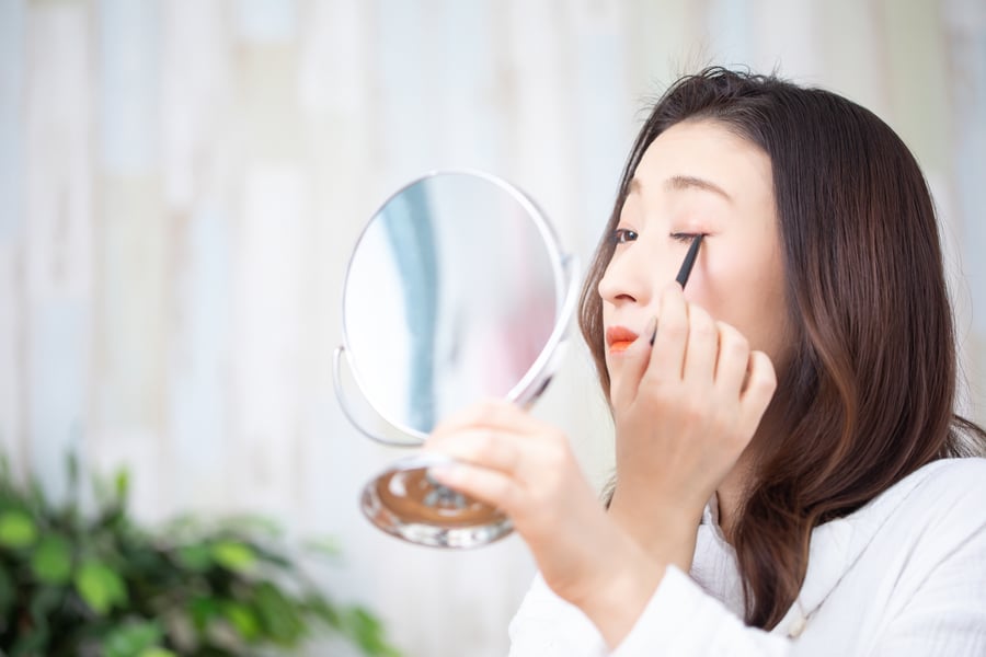 Người phụ nữ đang trang điểm mắt bằng bút kẻ mắt, nhìn vào gương tròn, nền là tường màu sáng.