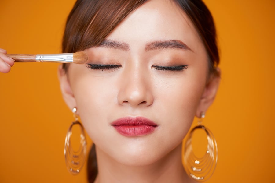 Người phụ nữ với đôi mắt nhắm, đang được trang điểm bằng cọ trên nền cam, đeo hoa tai lớn.