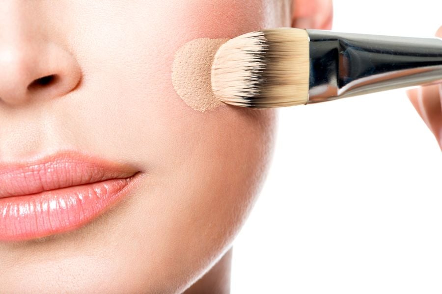 Cận cảnh một người đang dùng cọ trang điểm thoa phấn nền lên má.