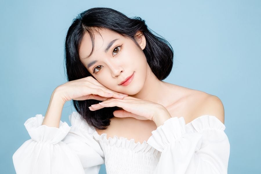 Một người mặc áo trắng tạo dáng với tay dưới cằm, nền xanh nhạt.