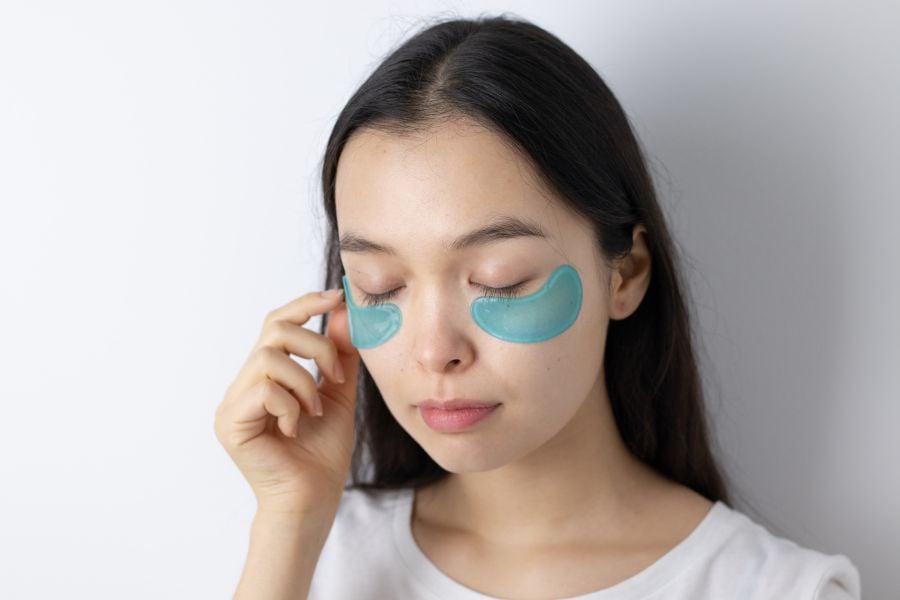 Người đang đắp mặt nạ mắt màu xanh dưới mắt, nhắm mắt thư giãn trong phòng sáng.