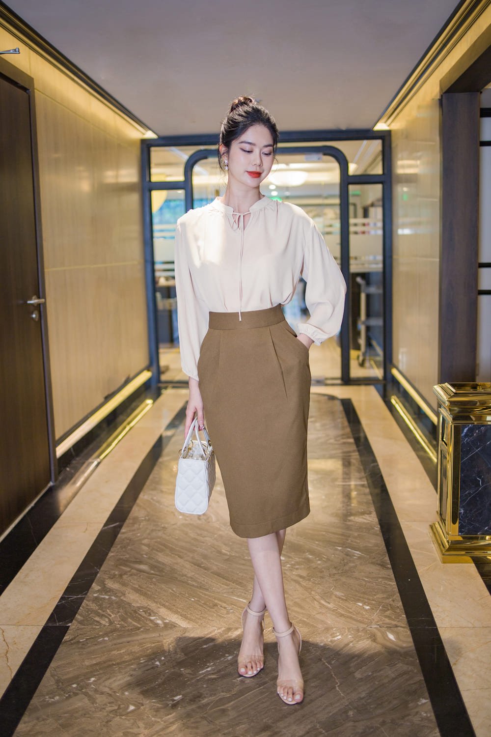 Người phụ nữ mặc áo trắng và váy nâu, đứng trong hành lang với túi xách màu trắng.