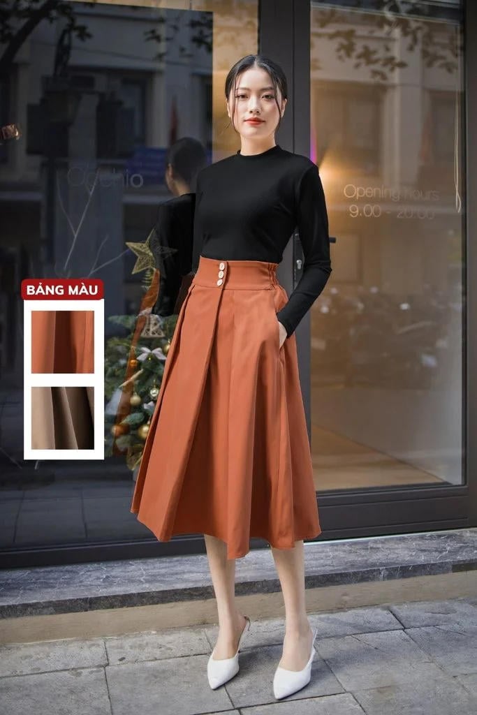 Người mẫu mặc áo đen và váy dài màu nâu cam đứng trước cửa kính, bên cạnh là bảng màu trang phục.