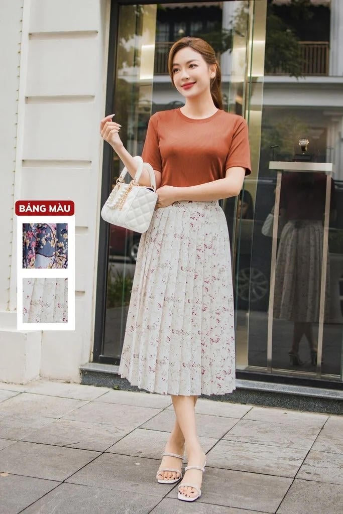 Người phụ nữ đứng trước cửa kính, mặc áo thun nâu, váy hoa dài, cầm túi xách trắng, mang dép quai mảnh.