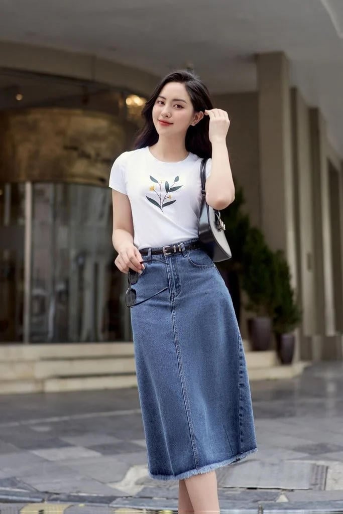 Người phụ nữ mặc áo trắng và váy jean đứng trước tòa nhà, tay giữ tóc, cười nhẹ nhàng.
