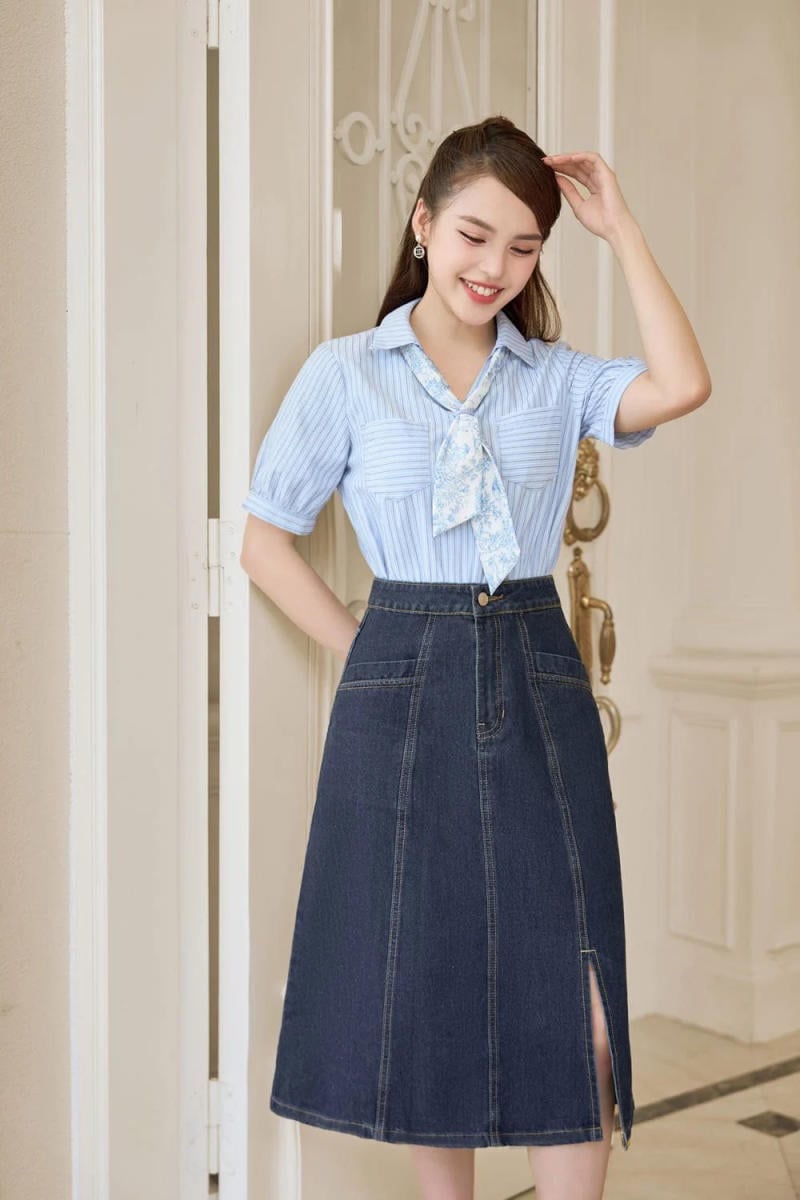 Người phụ nữ mặc áo sơ mi xanh và váy jean, đứng trước cửa, cười nhẹ và chỉnh tóc.
