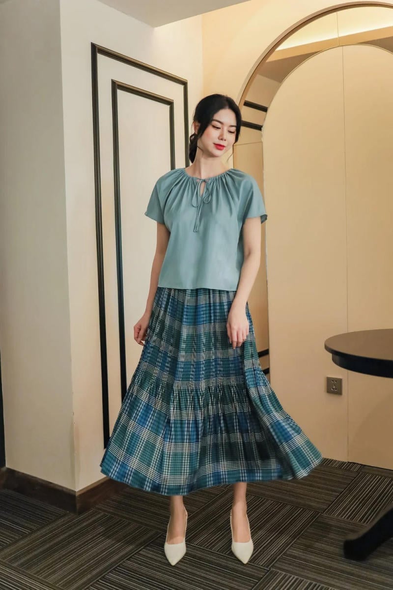 Người phụ nữ mặc áo xanh nhạt và váy kẻ caro xanh, đứng trong phòng có nền tường và cửa sổ trang trí.