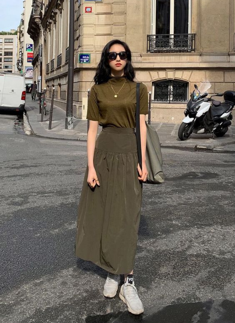 Người đang đứng trên đường phố, mặc áo thun và váy dài, đeo kính mát, có xe máy và tòa nhà phía sau.