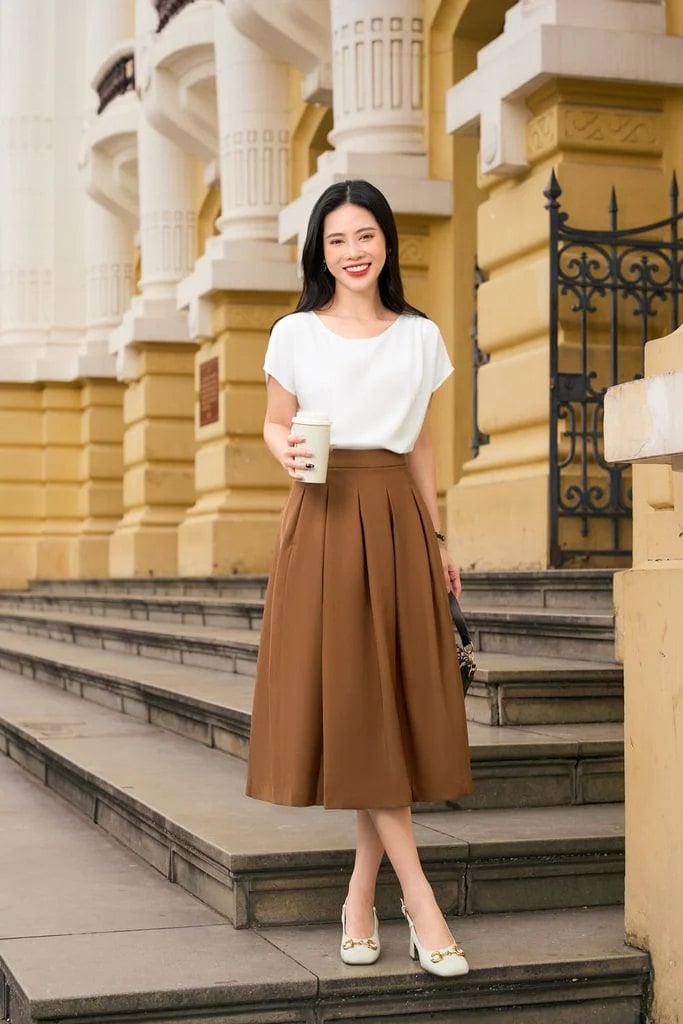 Người phụ nữ mặc váy nâu và áo trắng đứng trên cầu thang, tay cầm ly nước, phía sau là tòa nhà cổ điển.