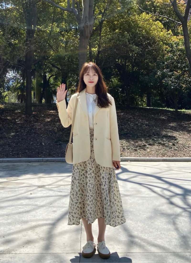 10 Cách Phối Đồ Với Chân Váy Hoa Nhí Giúp Bạn Tỏa Sáng Mùa Hè 27 Người phụ nữ đứng ngoài trời, mặc áo khoác màu kem và váy hoa, vẫy tay chào trước nền cây xanh.