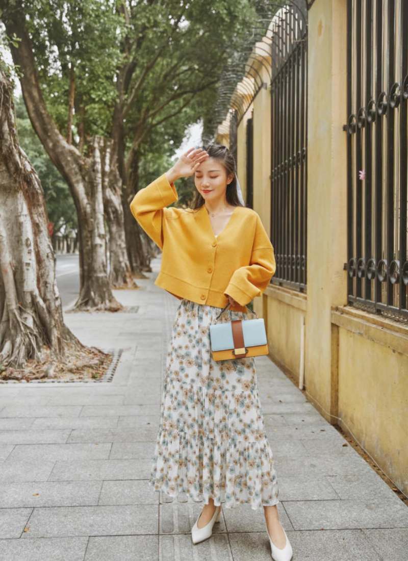 10 Cách Phối Đồ Với Chân Váy Hoa Nhí Giúp Bạn Tỏa Sáng Mùa Hè 21 Người phụ nữ mặc áo vàng, váy hoa, cầm túi xách đứng trên vỉa hè, cạnh hàng cây và hàng rào sắt.