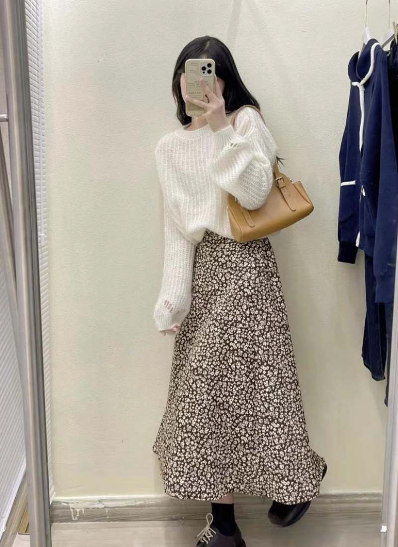 10 Cách Phối Đồ Với Chân Váy Hoa Nhí Giúp Bạn Tỏa Sáng Mùa Hè 22 Người mặc áo len trắng, váy hoa dài, cầm điện thoại trước gương trong phòng thử đồ, túi xách màu nâu bên vai.