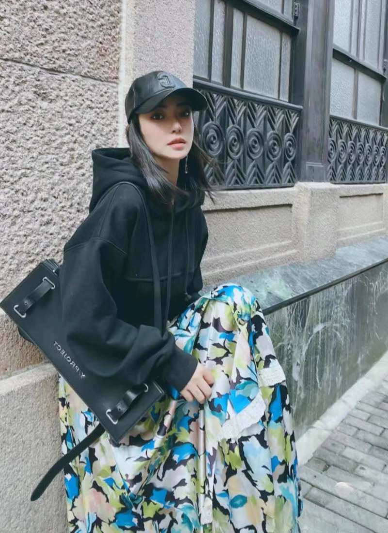 10 Cách Phối Đồ Với Chân Váy Hoa Nhí Giúp Bạn Tỏa Sáng Mùa Hè 24 Người mặc áo khoác đen và váy hoa, đội mũ lưỡi trai, ngồi cạnh bức tường có họa tiết trang trí.