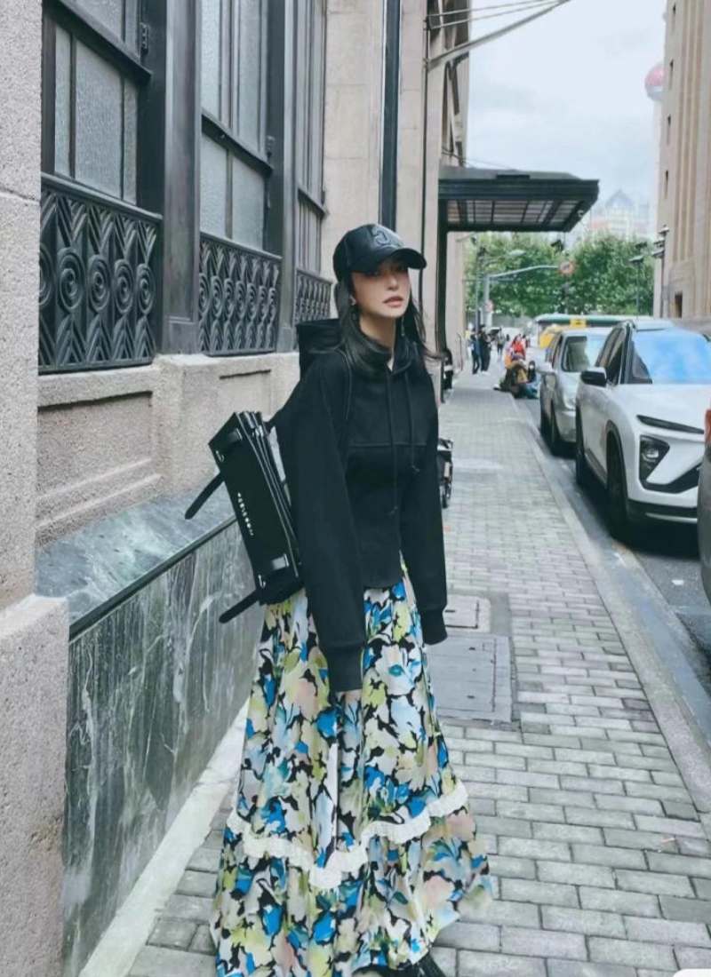 10 Cách Phối Đồ Với Chân Váy Hoa Nhí Giúp Bạn Tỏa Sáng Mùa Hè 25 Người mặc áo khoác đen, váy hoa dài và đội mũ lưỡi trai, đứng trên vỉa hè gần tòa nhà và xe cộ.