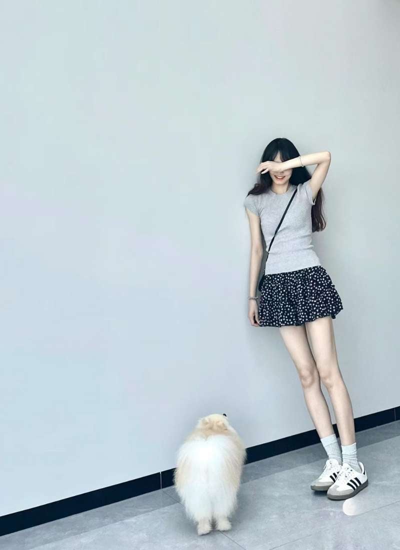 10 Cách Phối Đồ Với Chân Váy Hoa Nhí Giúp Bạn Tỏa Sáng Mùa Hè 11 Người mặc váy ngắn đứng cạnh chú chó lông xù, tay che mặt, trên nền tường màu xám nhạt.
