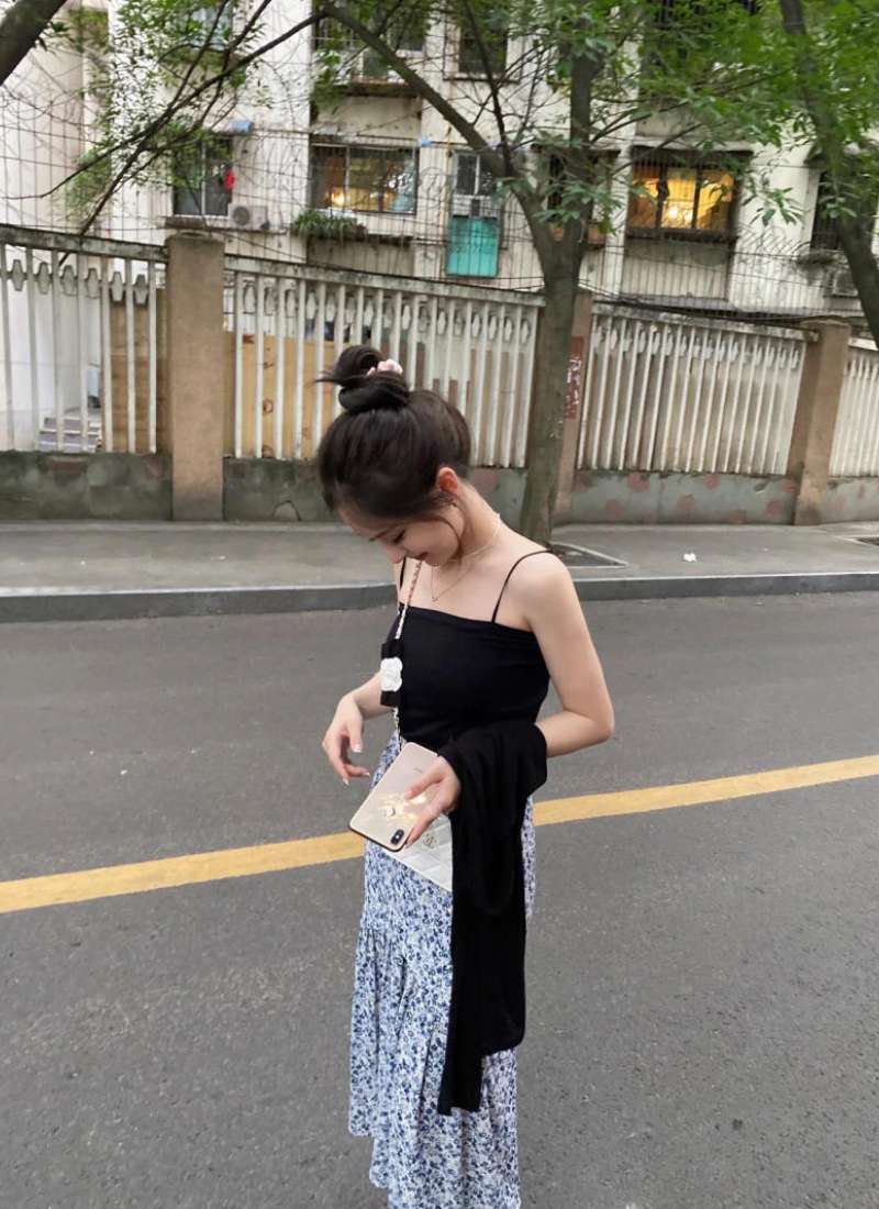 10 Cách Phối Đồ Với Chân Váy Hoa Nhí Giúp Bạn Tỏa Sáng Mùa Hè 16 Người phụ nữ đứng trên đường, mặc váy hoa và áo đen, cầm điện thoại và áo khoác, nền là hàng rào và cây xanh.