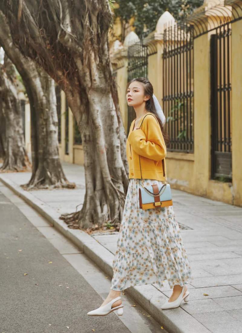 10 Cách Phối Đồ Với Chân Váy Hoa Nhí Giúp Bạn Tỏa Sáng Mùa Hè 20 Người phụ nữ mặc váy hoa và áo khoác vàng, đi bộ trên vỉa hè bên cạnh hàng cây cổ thụ.