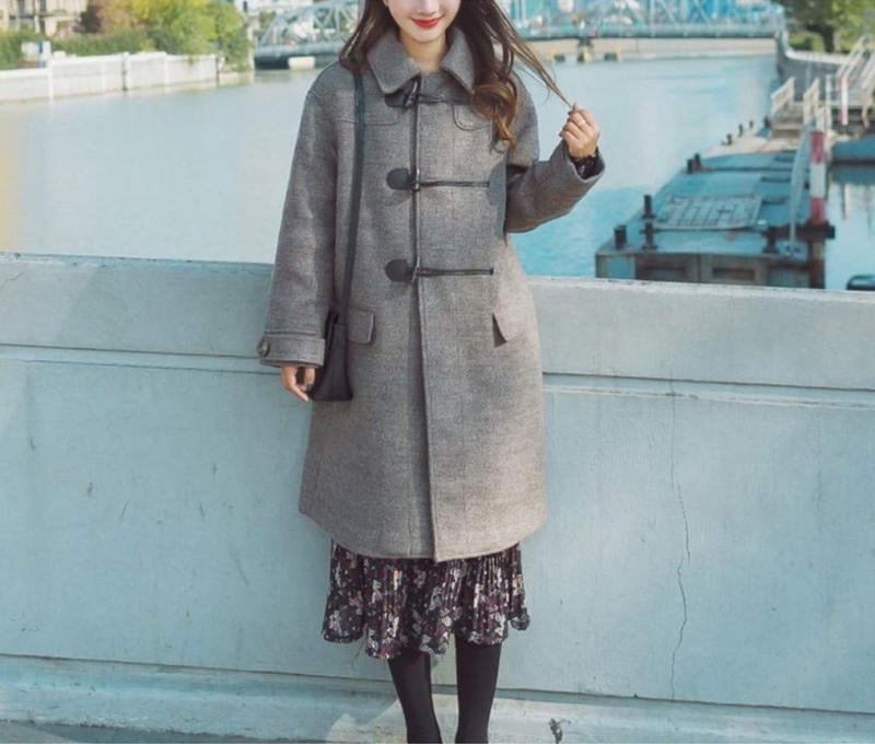 10 Cách Phối Đồ Với Chân Váy Hoa Nhí Giúp Bạn Tỏa Sáng Mùa Hè 28 Người mặc áo khoác xám đứng cạnh lan can, nền là dòng sông và cây cầu.