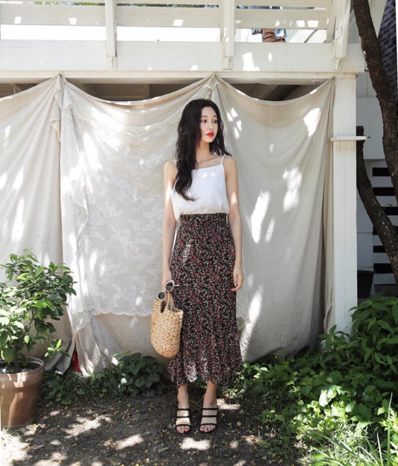 10 Cách Phối Đồ Với Chân Váy Hoa Nhí Giúp Bạn Tỏa Sáng Mùa Hè 15 Người phụ nữ đứng trong vườn, mặc áo trắng và váy hoa, cầm túi cói, phông nền là tấm vải trắng treo.