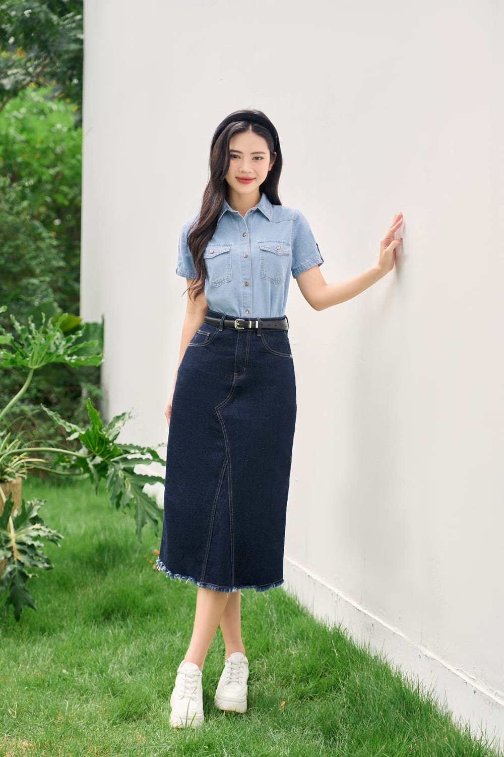 Người phụ nữ mặc áo sơ mi và váy jean, đứng dựa vào tường trắng, nền là cây xanh.