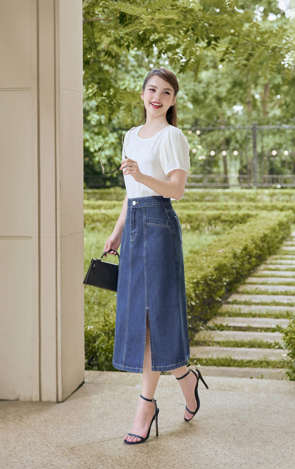 Người đang đứng ngoài trời, mặc áo trắng và váy denim dài, cầm kính mát và túi xách nhỏ, có hàng cây xanh phía sau.