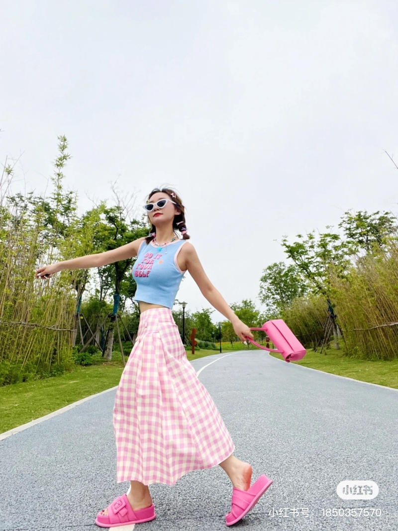 Người phụ nữ mặc áo xanh, váy kẻ caro, cầm túi hồng, đi dép hồng, đi bộ trên con đường trong công viên.