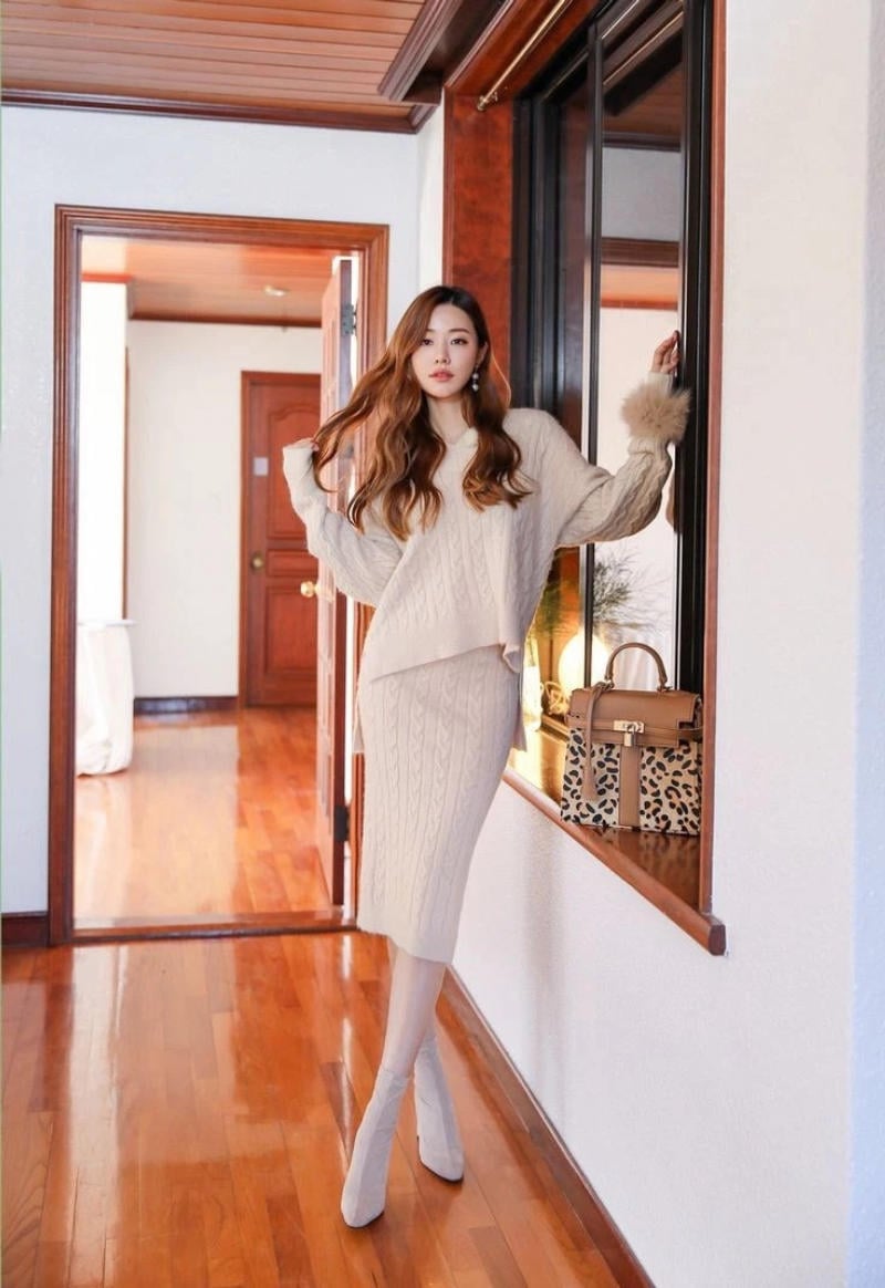 Người phụ nữ mặc trang phục len màu be đứng cạnh cửa sổ, cạnh đó là một túi xách họa tiết da báo.