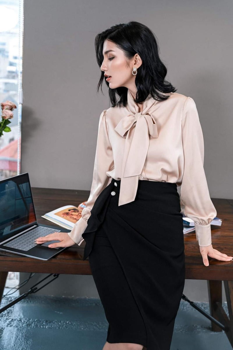 Người phụ nữ mặc áo sơ mi và váy, đứng tựa vào bàn làm việc với laptop và sách, nền văn phòng hiện đại.