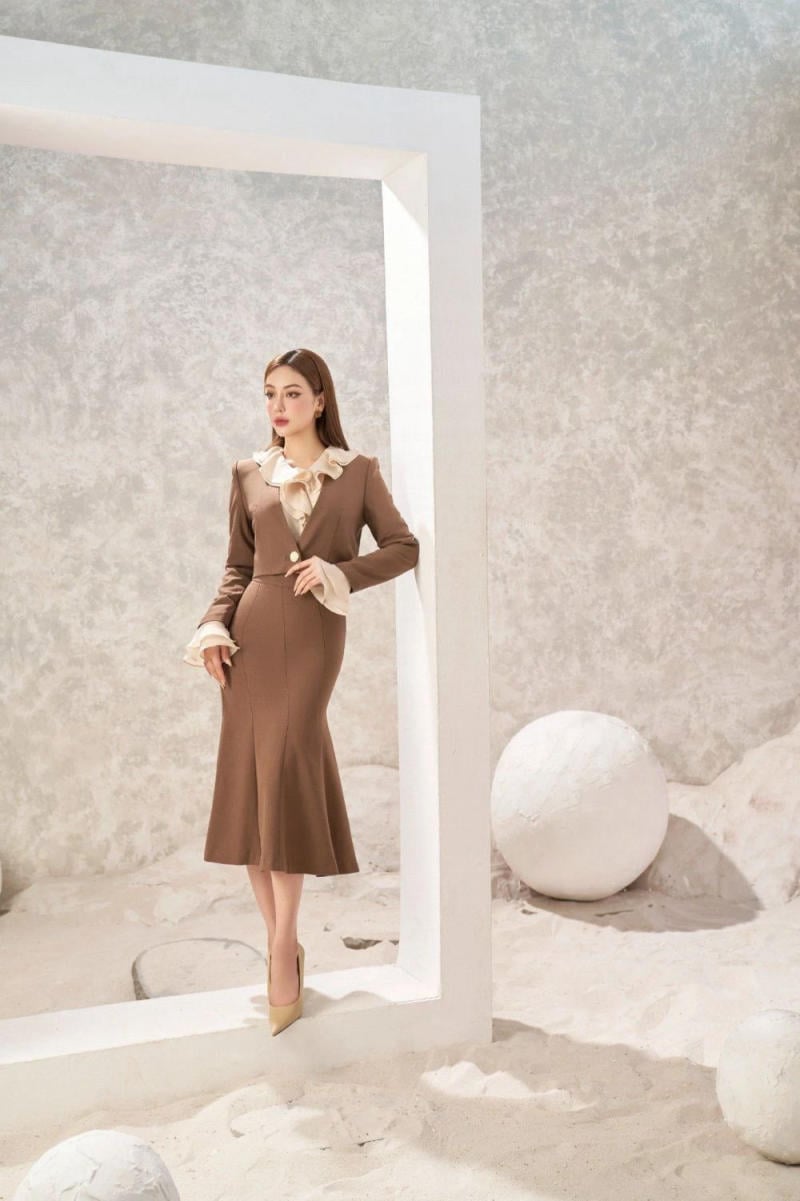 Người phụ nữ mặc váy nâu đứng dựa vào khung trắng trong không gian có nền cát và các quả cầu trắng.