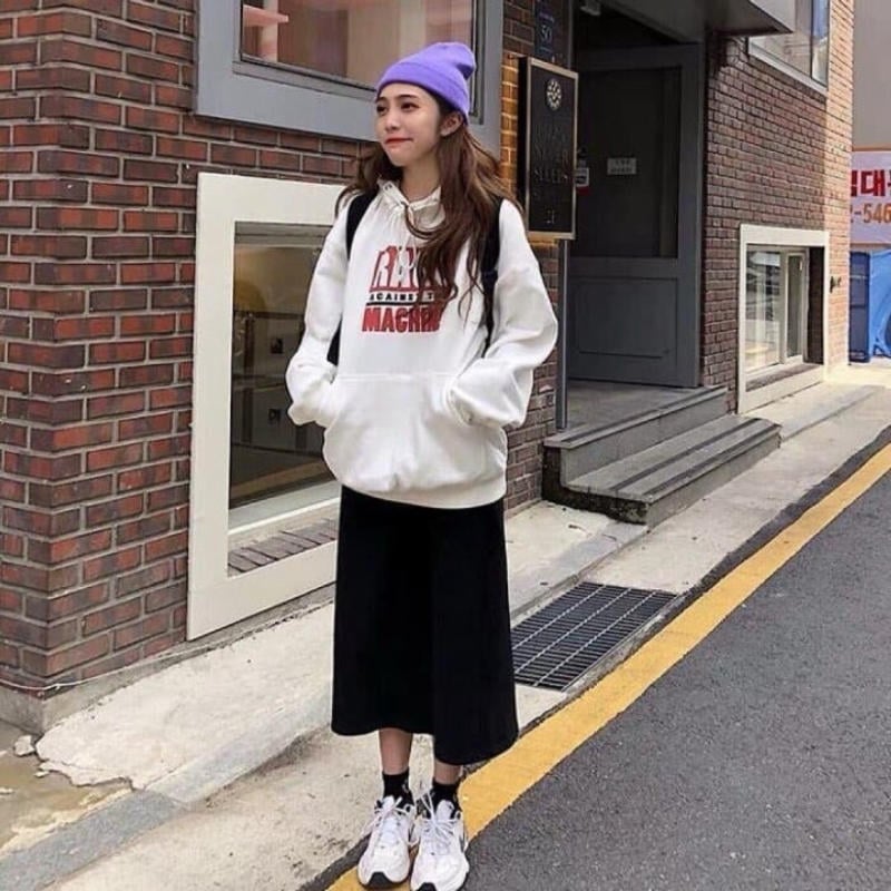 Người mặc áo hoodie trắng và mũ tím đứng trên vỉa hè, cạnh bức tường gạch.
