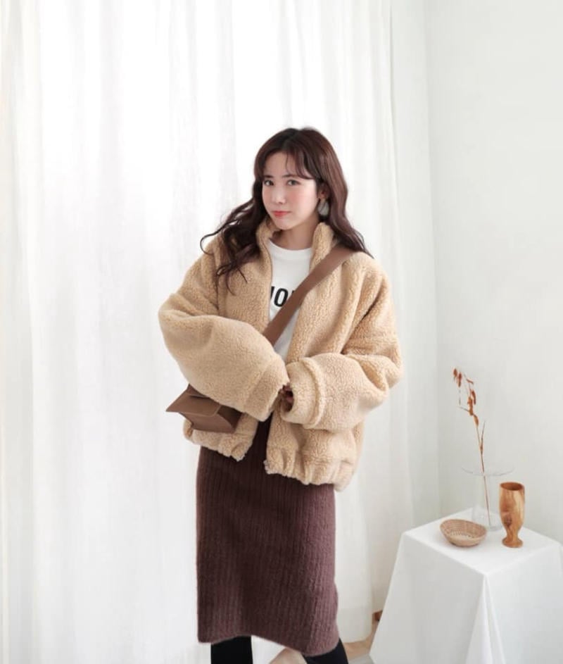 Người phụ nữ mặc áo khoác lông màu be và váy nâu đứng trước phông nền trắng trong phòng sáng.