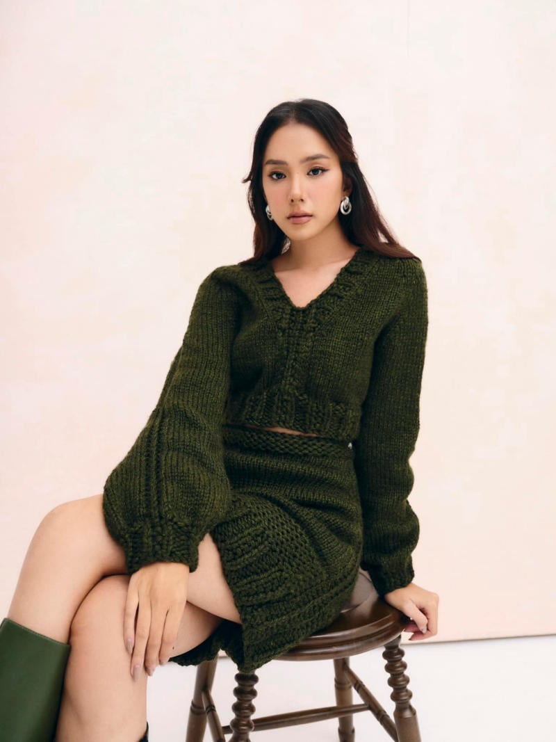 Người phụ nữ ngồi trên ghế, mặc trang phục len màu xanh lá, nền trắng.