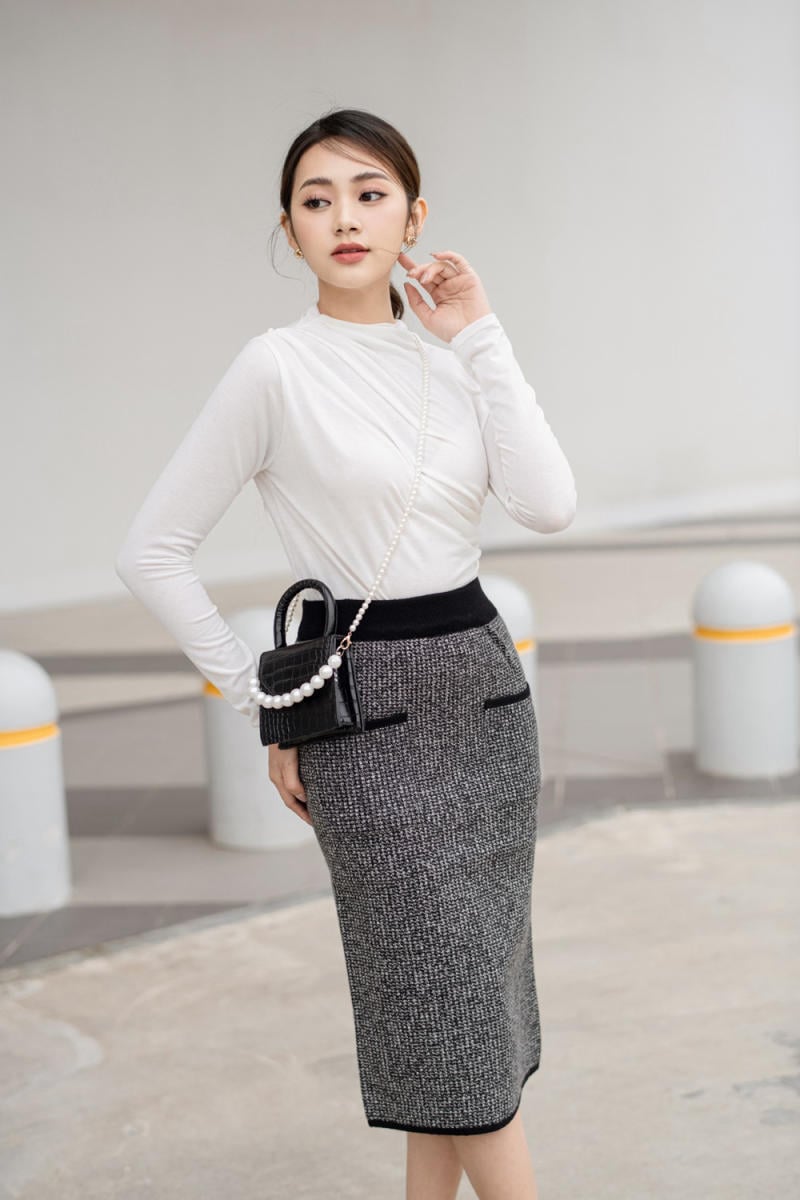 Người mặc áo trắng và chân váy xám, đeo túi xách nhỏ màu đen, đứng trên nền bê tông.