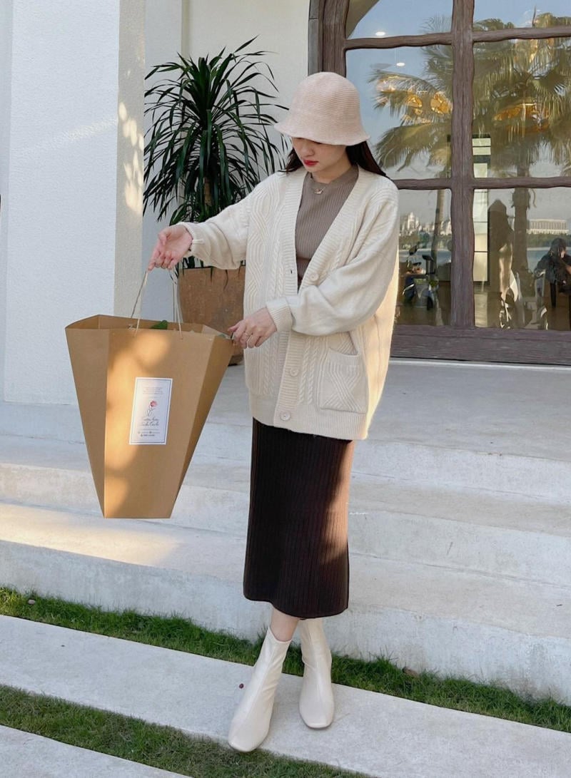 Người phụ nữ đội mũ, mặc áo cardigan đứng trên bậc thềm cầm túi giấy lớn, nền là cửa sổ và cây xanh.