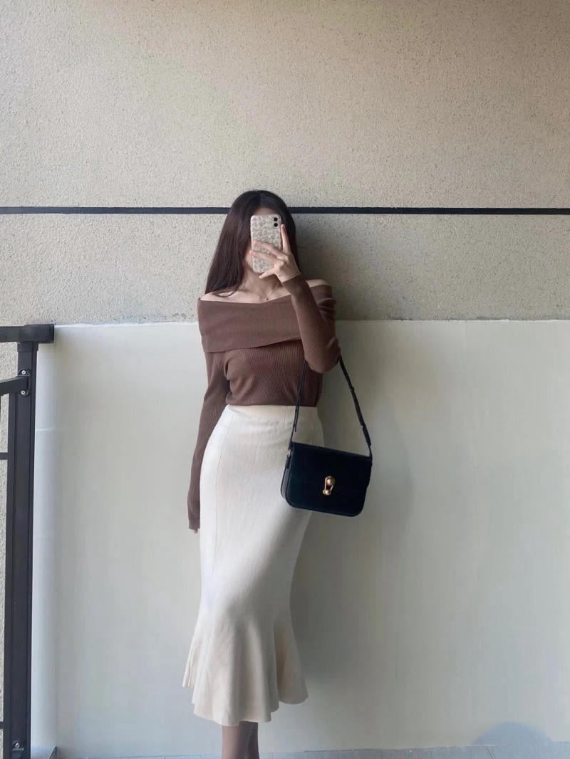 Người phụ nữ mặc áo trễ vai nâu, váy trắng, cầm điện thoại chụp ảnh, đứng cạnh tường và lan can.