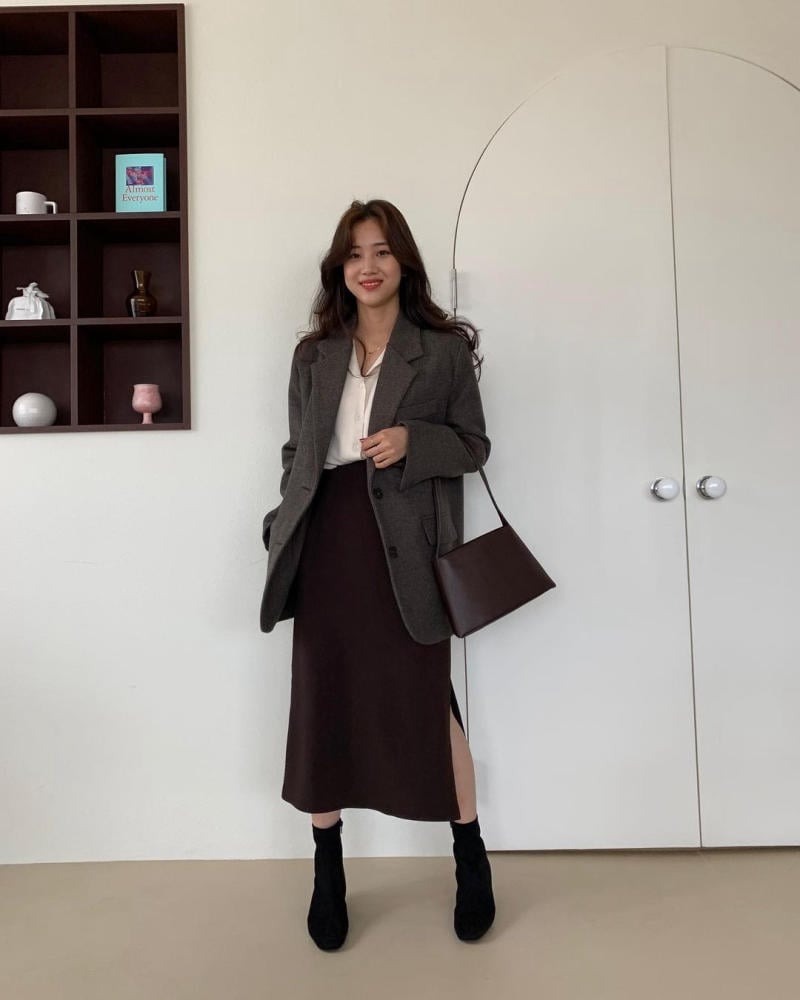 Người phụ nữ mặc áo blazer và chân váy dài, đứng trước cửa, bên cạnh kệ gỗ có vài đồ trang trí.