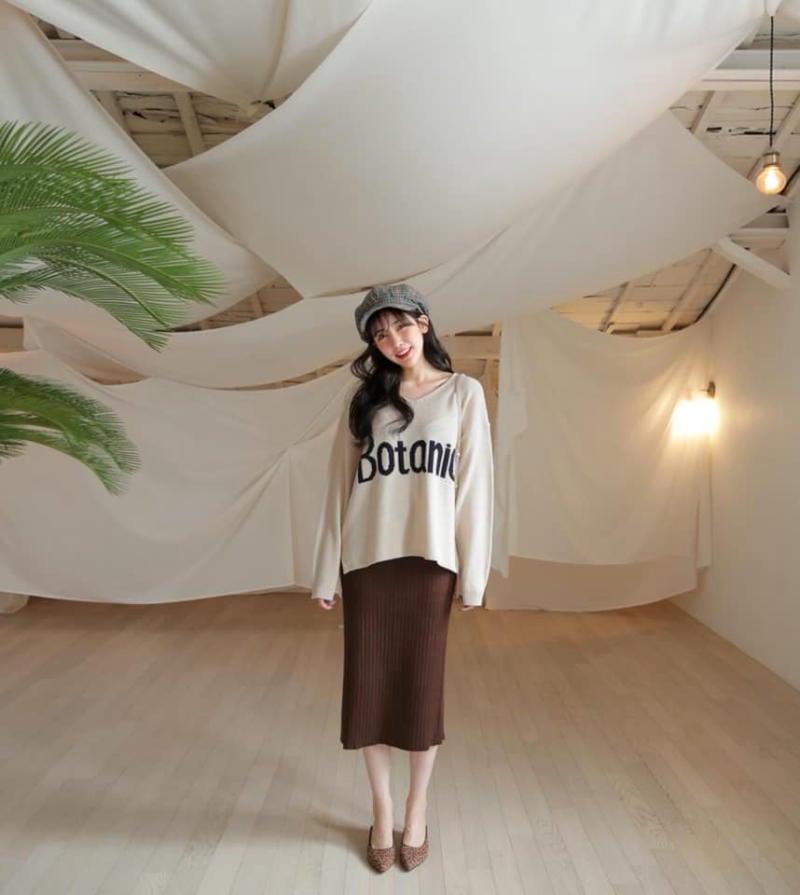 Người phụ nữ trong trang phục thời trang đứng trong căn phòng sáng, trần trang trí vải trắng, gần cây xanh.