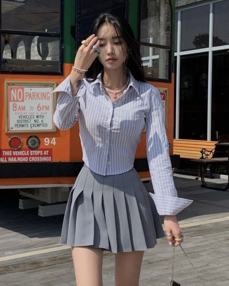 Người mặc áo sơ mi kẻ sọc và váy xếp ly đứng trước xe buýt màu cam, tay cầm điện thoại.