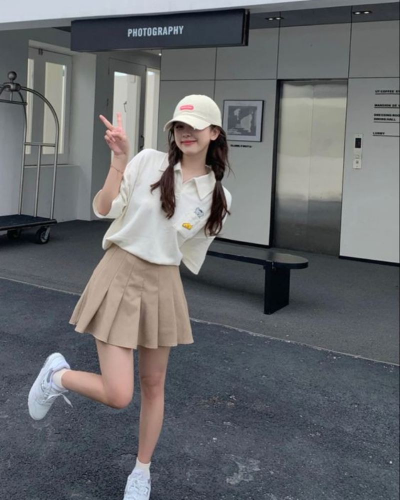 Người mặc váy ngắn và áo thun, đội mũ lưỡi trai, tạo dáng vui vẻ trước tòa nhà.