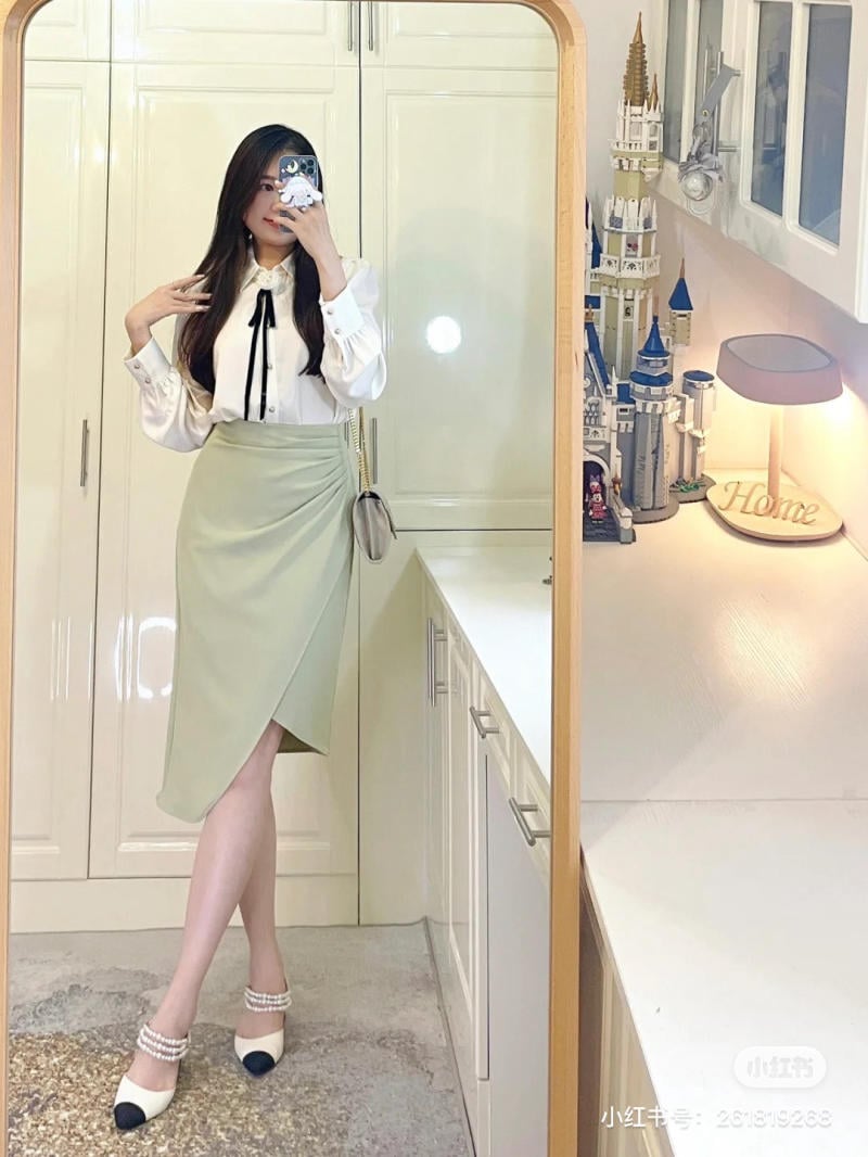 Người chụp gương trong phòng, mặc áo trắng, váy xanh nhạt, bên cạnh là mô hình lâu đài và đèn bàn.