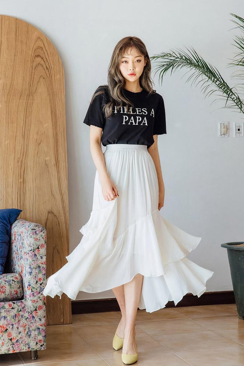 Người mặc áo thun đen và váy trắng đứng trong phòng, bên cạnh ghế hoa và cây cảnh.