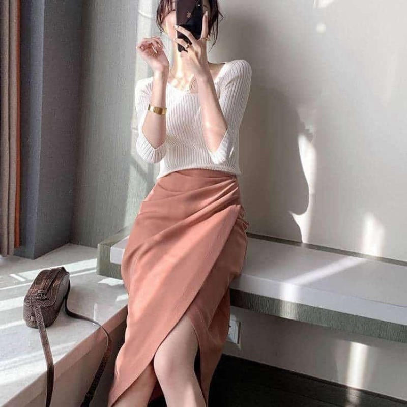 Người phụ nữ mặc áo trắng và váy dài màu cam đang ngồi trên ghế, chụp ảnh tự sướng bằng điện thoại.
