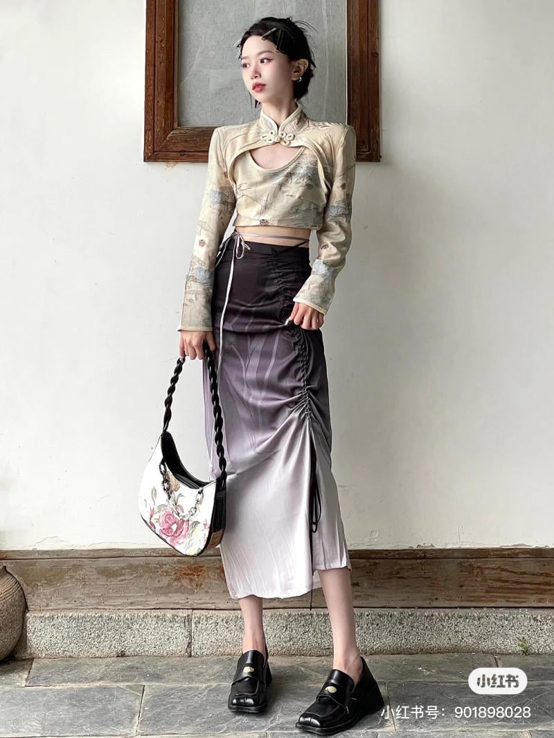 Người mẫu đứng cạnh bức tường, mặc áo kiểu truyền thống và váy dài, cầm túi xách, đi giày đen.