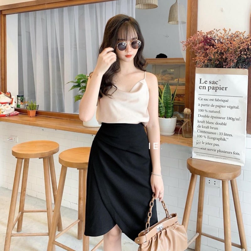 Người phụ nữ đeo kính râm đứng cạnh quầy bar với ghế gỗ, đằng sau là cây xanh và túi giấy.