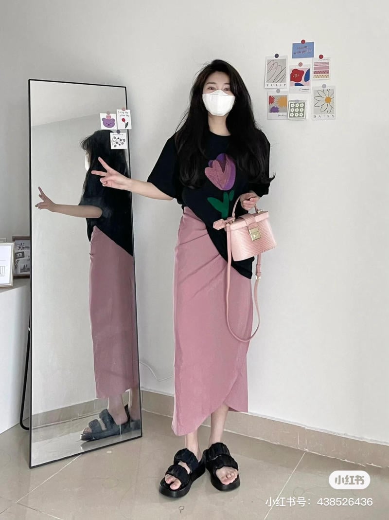 Người đeo khẩu trang, mặc áo in hoa, chân váy hồng, giày đen, cầm túi xách, đứng trước gương và tường trang trí.