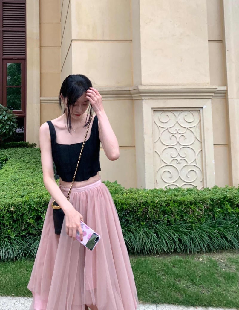 Người mặc áo đen và váy hồng đứng trước tòa nhà, tay giữ điện thoại với ốp lưng hoa.